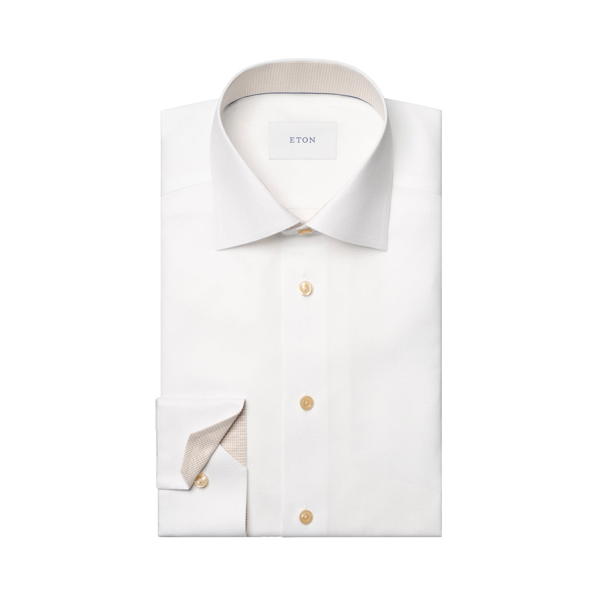A folded white dress shirt with a spread collar and a single cuff turned up, showing a light beige inner lining. The shirt has gold buttons and a visible neck label that reads ETON. Eton Shirt available at Sakks Menswear in Southampton.