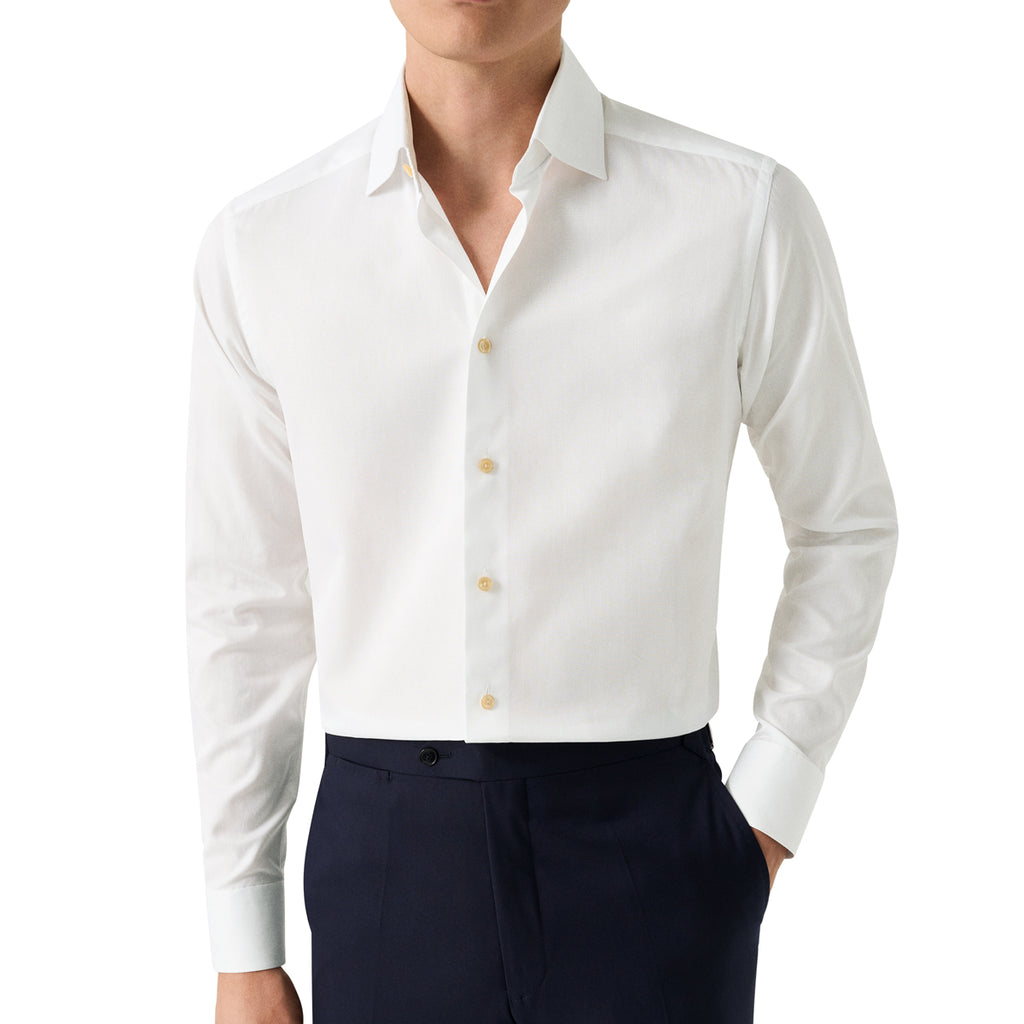 A man wearing a crisp white long-sleeve button-down dress shirt with light-coloured buttons, paired with dark navy trousers, shown from the chin down on a plain white background. Eton Shirt available at Sakks Menswear in Southampton.