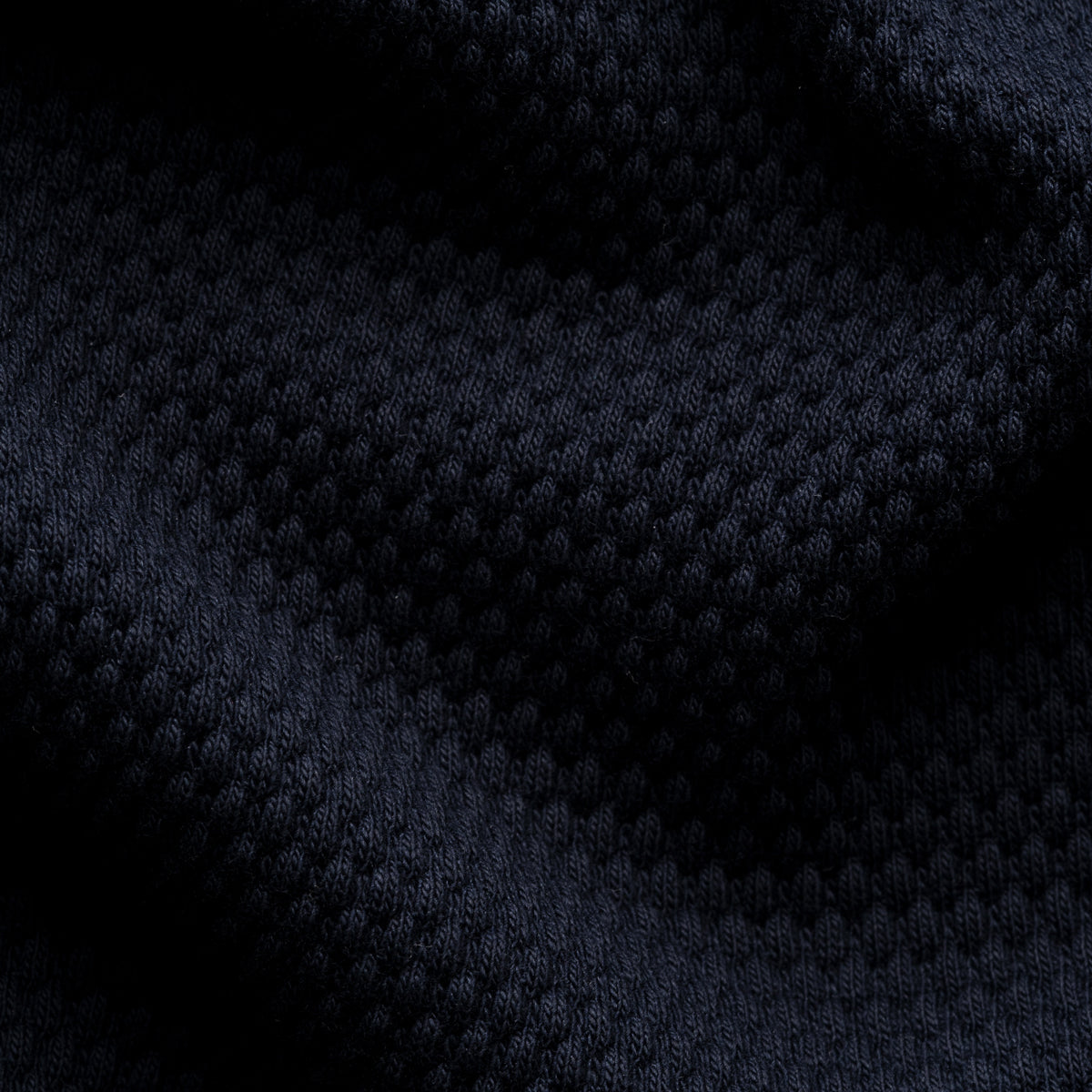 A close-up of dark navy blue textured knit fabric with a waffle or dimple pattern, shown with soft folds and shadows. Eton Polo available at Sakks Menswear in Southampton.