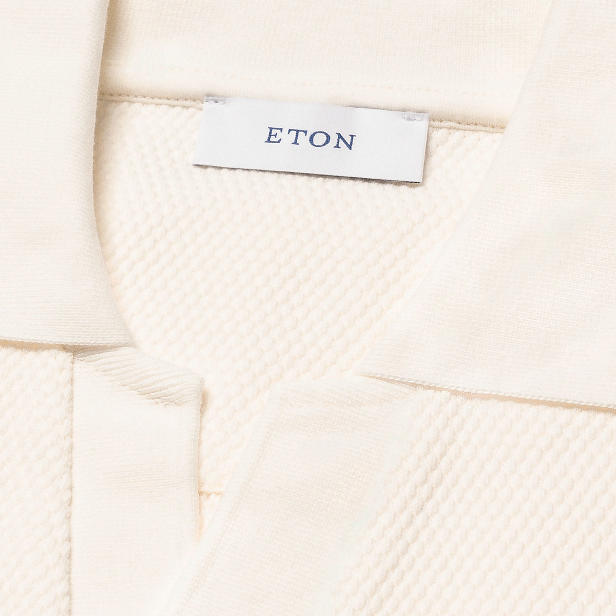 Close-up of a cream-colored textured knit garment, showing a white brand label with the name ETON printed in navy blue. Eton Polo available at Sakks Menswear in Southampton.
