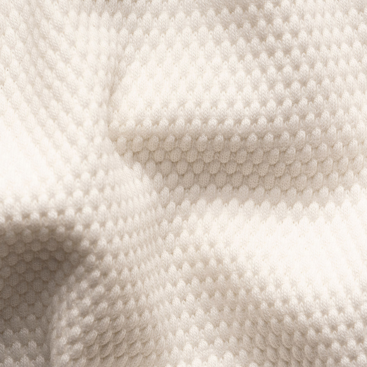A close-up of a textured knit fabric in a warm off-white or cream color, showing the detailed looped knit pattern and soft folds. Eton Polo available at Sakks Menswear in Southampton.