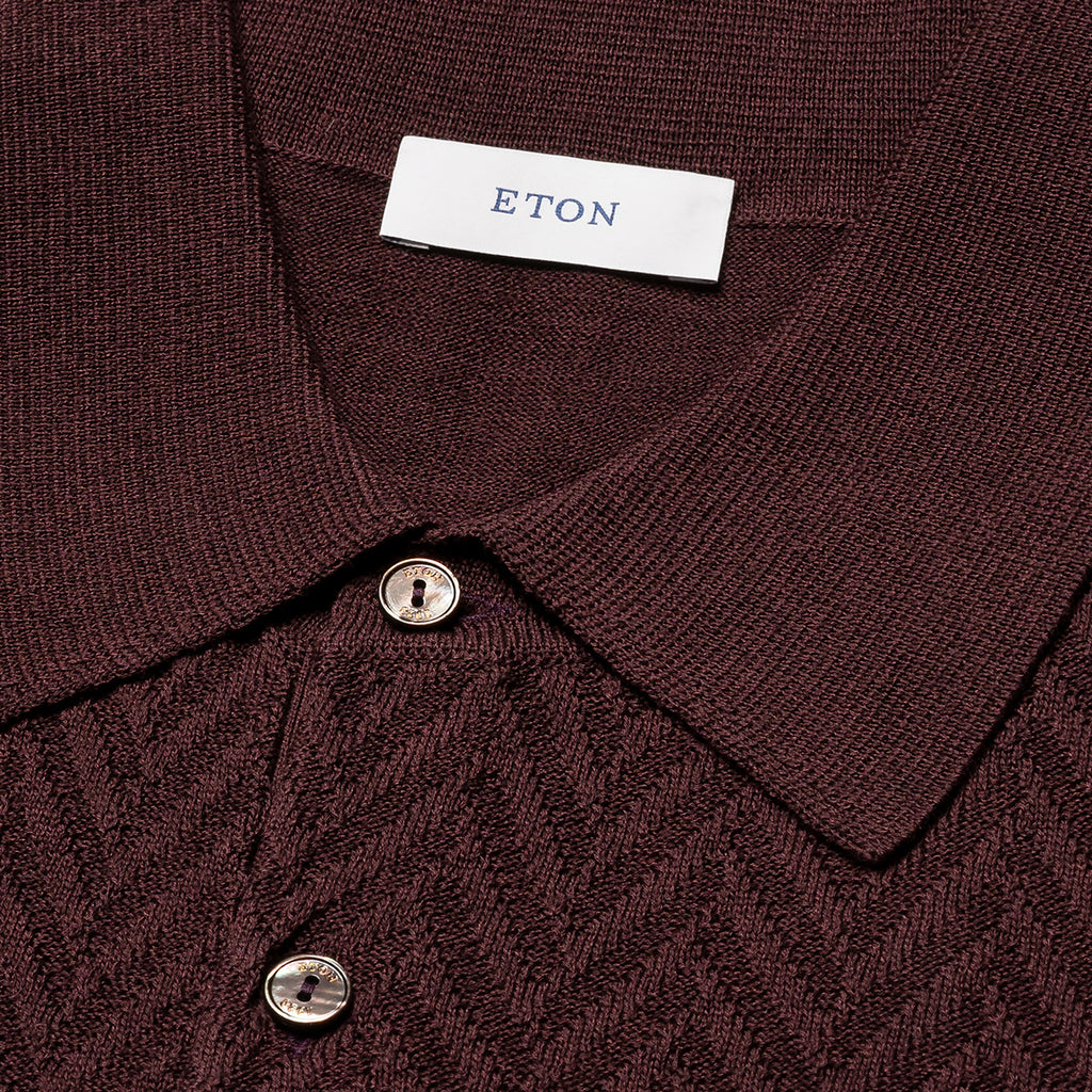 Close-up of a dark burgundy knit polo shirt with a textured herringbone pattern, featuring a white neck label with the brand name 'ETON' and two silver buttons. Eton Polo. Sakks Menswear Southampton.