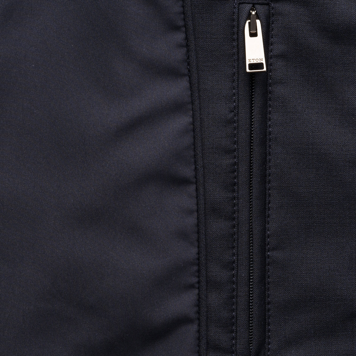 Close-up of a dark navy jacket or fabric panel with a vertical silver zipper and a small visible 'ETON' brand tab on the zipper pull. Eton Gilet. Sakks Menswear Southampton.