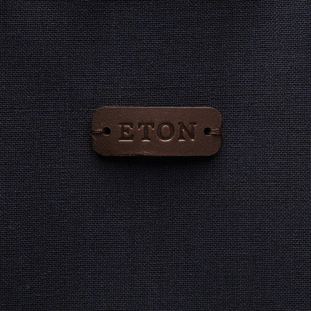 A close-up of a dark brown leather brand patch embossed with the word 'ETON' on a dark navy textured fabric background. Eton Gilet. Sakks Menswear Southampton.
