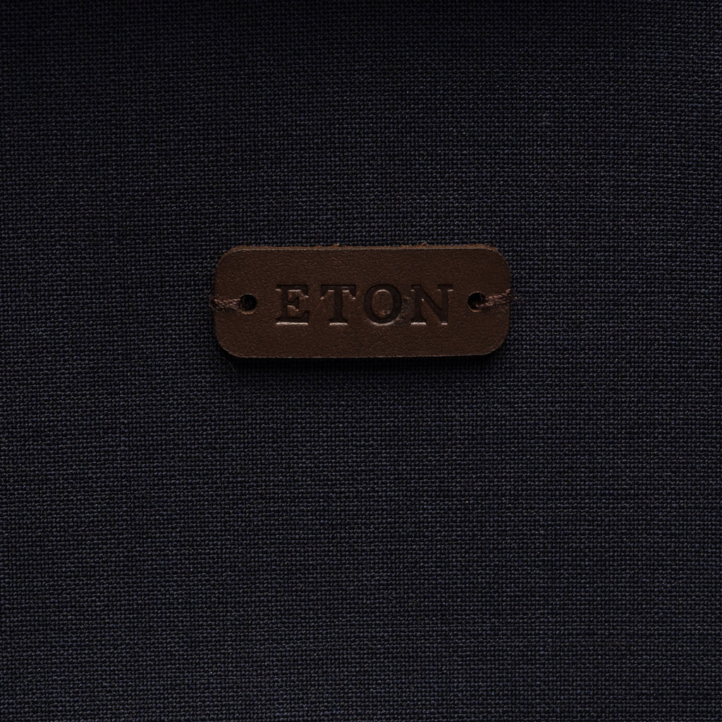 A close-up of a dark brown leather patch embossed with the brand name 'ETON' on a dark navy blue textured fabric background. Eton Shirt Jacket. Sakks Menswear Southampton.