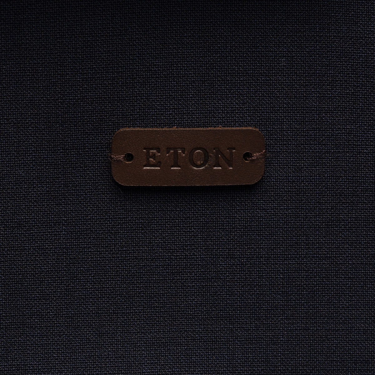 A close-up of a dark brown leather patch embossed with the brand name 'ETON' on a dark navy blue textured fabric background. Eton Shirt Jacket. Sakks Menswear Southampton.