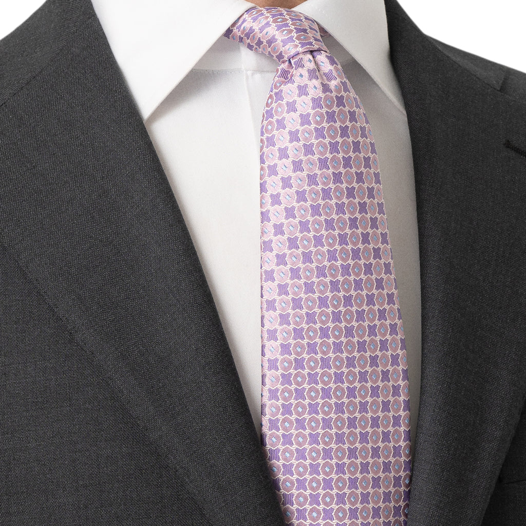 A close-up of a men's necktie with a lavender and beige geometric pattern, styled on a white dress shirt and dark gray suit jacket. Eton Ties available at Sakks Menswear in Southampton.