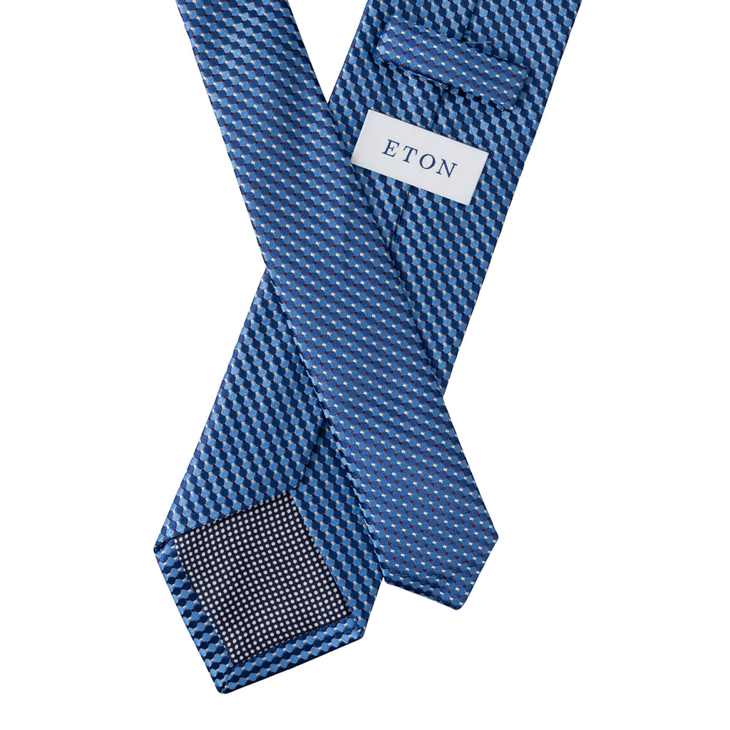 A blue patterned necktie with a small white label reading ETON, shown crossed over itself on a plain white background. Eton Ties available at Sakks Menswear in Southampton.