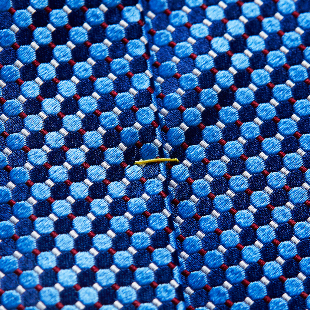 Close-up of a textured woven necktie fabric with a repeating geometric pattern of light and medium blue circles, dark navy hexagons, and thin red lines, featuring a small yellow tie clip on the front. Eton Ties available at Sakks Menswear in Southampton.