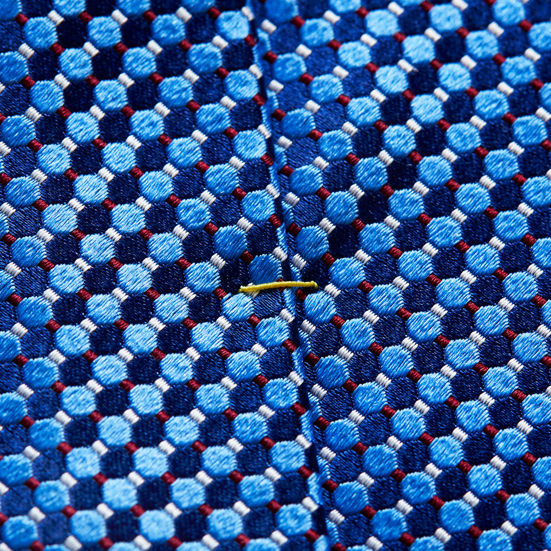 Close-up of a textured woven necktie fabric with a repeating geometric pattern of light and medium blue circles, dark navy hexagons, and thin red lines, featuring a small yellow tie clip on the front. Eton Ties available at Sakks Menswear in Southampton.