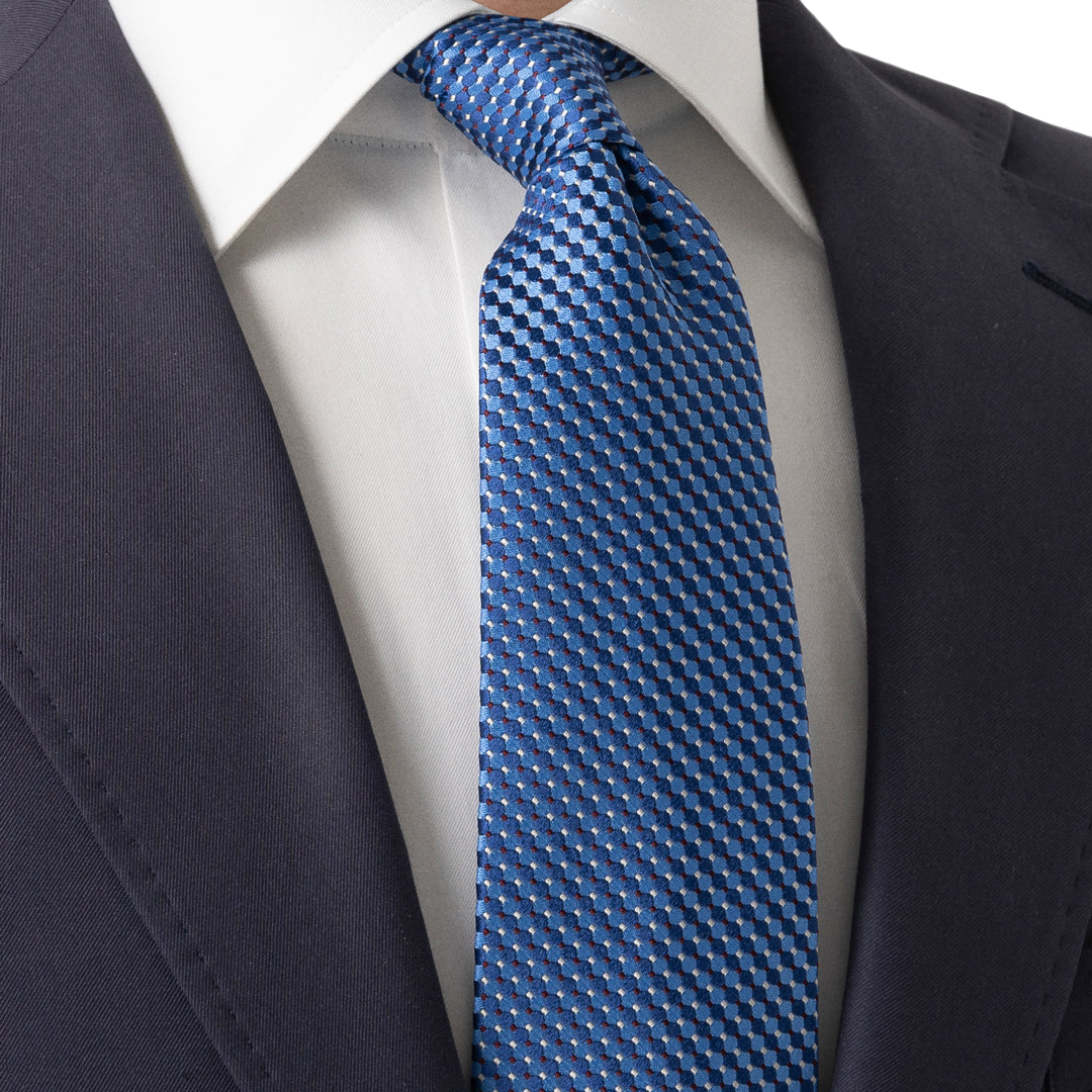 A close-up of a men's necktie with a blue and gray geometric pattern, styled on a white dress shirt and dark navy suit jacket. Eton Ties available at Sakks Menswear in Southampton.