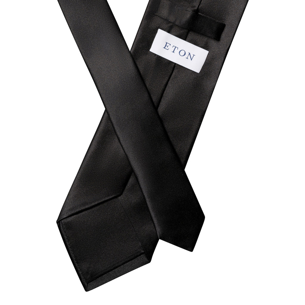 A black satin necktie with a white rectangular label reading 'ETON' in navy blue, shown crossed over itself on a plain white background. Eton Ties available at Sakks Menswear in Southampton.