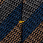 Close-up of a textured woven fabric with a geometric pattern in dark gray, navy blue, and brown, featuring a small yellow embroidered accent near the center. Eton Ties available at Sakks Menswear in Southampton.