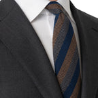 A close-up of a men's necktie with diagonal stripes of navy blue, brown, and beige, styled with a white dress shirt and a dark gray suit jacket. Eton Ties available at Sakks Menswear in Southampton.