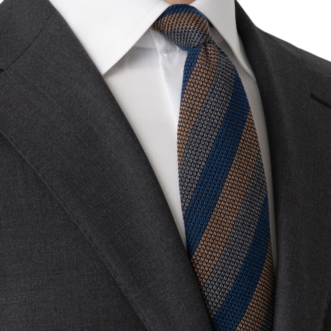 A close-up of a men's necktie with diagonal stripes of navy blue, brown, and beige, styled with a white dress shirt and a dark gray suit jacket. Eton Ties available at Sakks Menswear in Southampton.