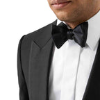 A close-up of a man wearing a classic black tuxedo jacket with satin peak lapels, a crisp white dress shirt, and a matching black satin bow tie, against a plain white background. Eton Bow Ties available at Sakks Menswear in Southampton.