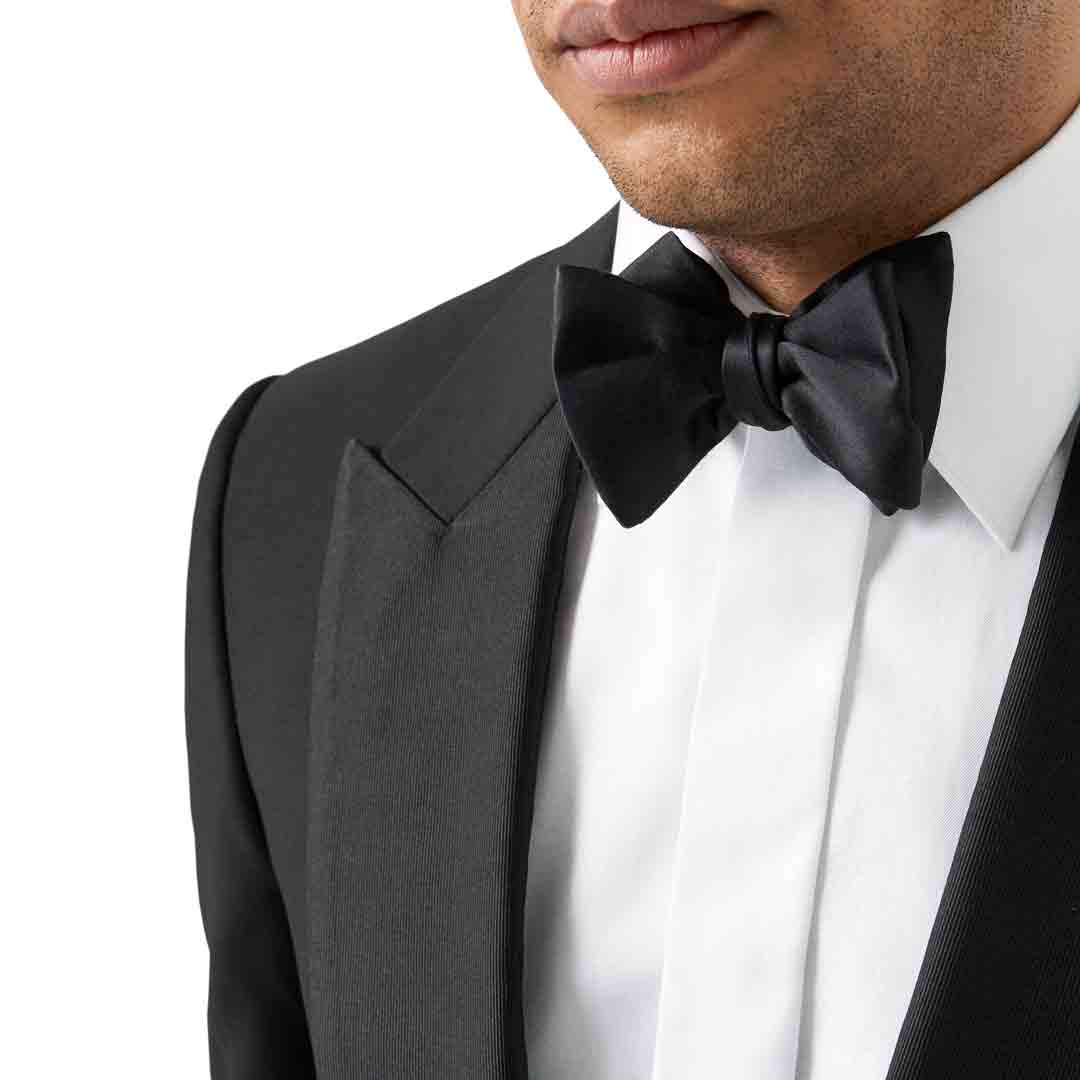 A close-up of a man wearing a classic black tuxedo jacket with satin peak lapels, a crisp white dress shirt, and a matching black satin bow tie, against a plain white background. Eton Bow Ties available at Sakks Menswear in Southampton.