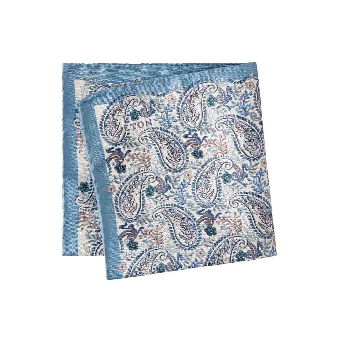 A folded square silk pocket square featuring a blue and beige paisley pattern with a solid light blue border, displayed on a clean white background. Eton Pocket Square available at Sakks Menswear in Southampton.