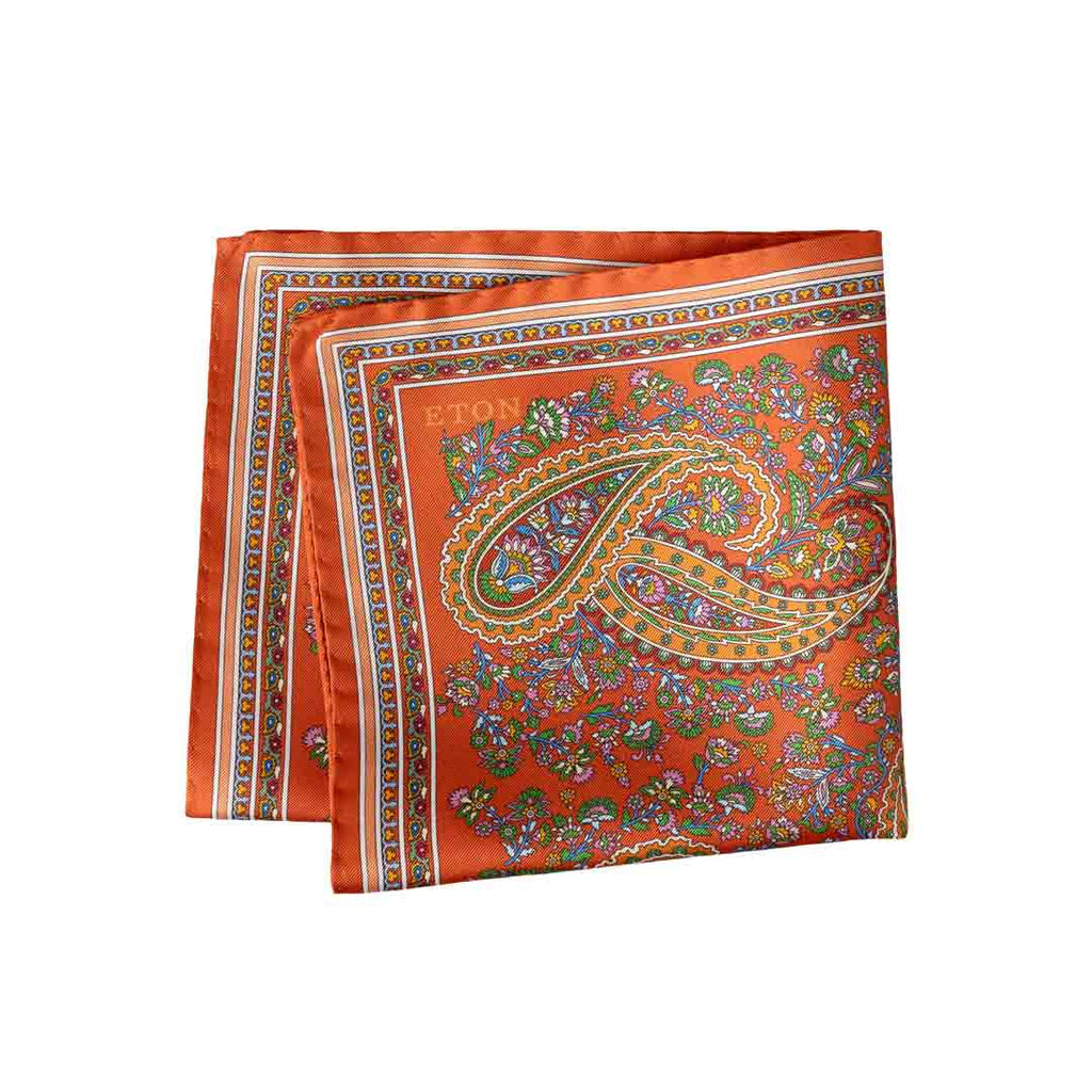 An orange silk scarf with a detailed paisley pattern in green, gold, and blue, featuring the brand name 'ETON' printed in gold near the top left corner. Eton Pocket Square available at Sakks Menswear in Southampton.
