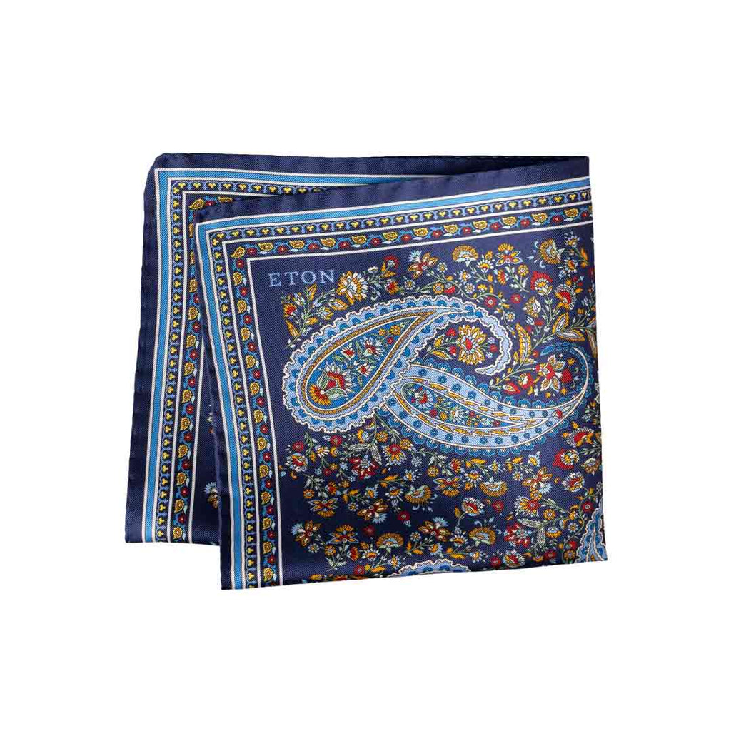 A folded navy blue silk-like pocket square with a detailed paisley and floral pattern in gold, blue, red, and white. The brand name 'ETON' is printed in white near the top left corner of the design. Eton Pocket Square available at Sakks Menswear in Southampton.
