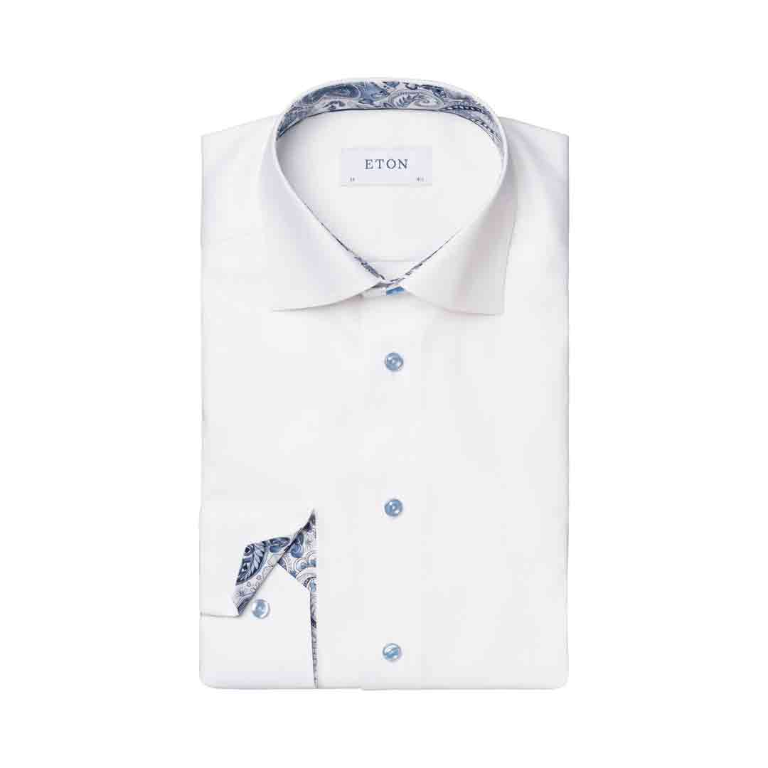A folded white dress shirt with a blue paisley patterned inner collar and matching pocket square, featuring light blue buttons and the brand name ETON on the collar label. Eton Shirt available at Sakks Menswear in Southampton.