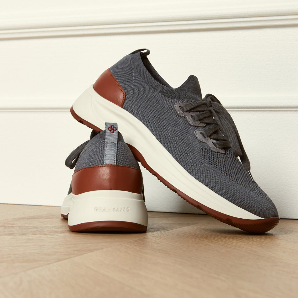 A pair of low-top athletic sneakers in dark grey mesh with brown leather accents and thick off-white midsoles, photographed on a light wood floor against a white panelled wall. Gran Sasso Sneakers available at Sakks Menswear in Southampton.