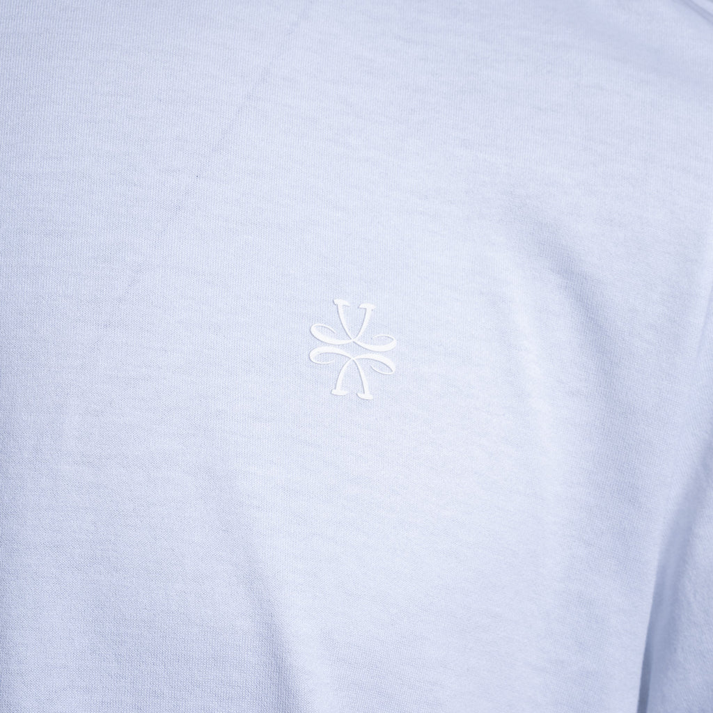 A close-up of a light periwinkle blue heathered fabric, likely a t-shirt, featuring a small white embroidered stylized logo in the center of the chest. Jacob Cohën T-Shirt available at Sakks Menswear in Southampton.