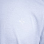 A close-up of a light periwinkle blue heathered fabric, likely a t-shirt, featuring a small white embroidered stylized logo in the center of the chest. Jacob Cohën T-Shirt available at Sakks Menswear in Southampton.