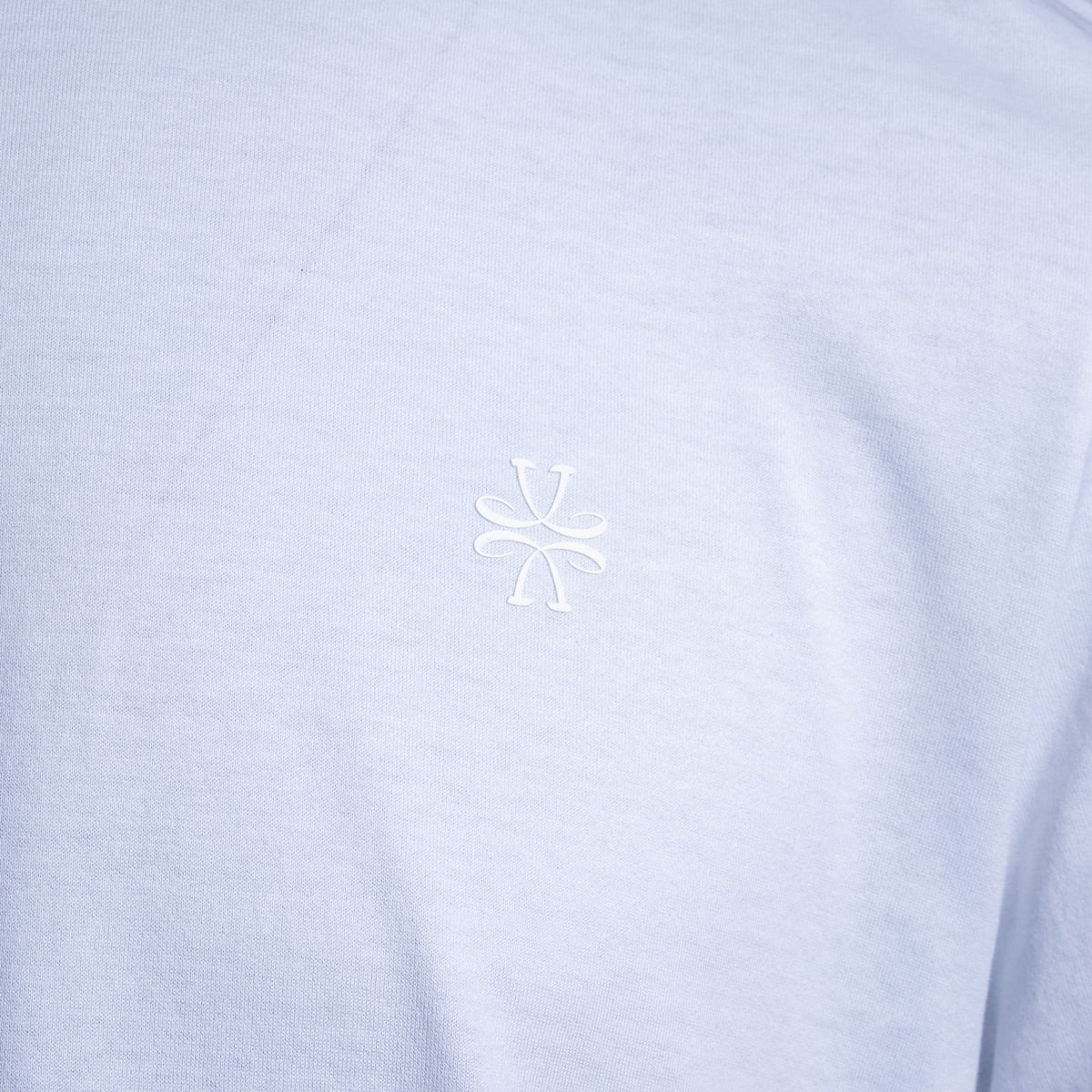 A close-up of a light periwinkle blue heathered fabric, likely a t-shirt, featuring a small white embroidered stylized logo in the center of the chest. Jacob Cohën T-Shirt available at Sakks Menswear in Southampton.