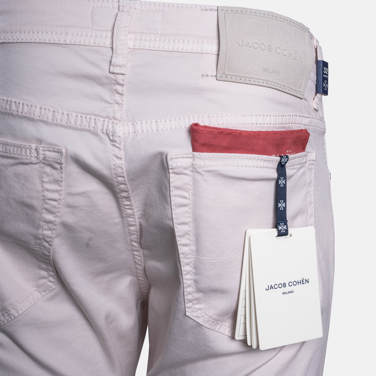 Close-up of the back waistband and right pocket of light pink denim jeans by Jacob Cohen, showing a red pocket lining, a dark blue brand tab, and a white hangtag. Jacob Cohën Chinos available at Sakks Menswear in Southampton.