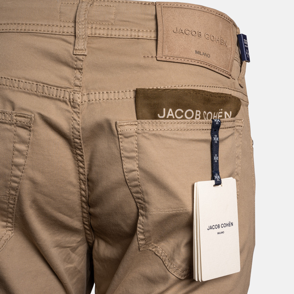 Close-up of the back waistband and pocket area of tan denim jeans by Jacob Cohen, showing the back patch, brown brand tab, hanging white product tag, and blue brand ribbon on a light gray background. Jacob Cohën Chinos available at Sakks Menswear in Southampton.