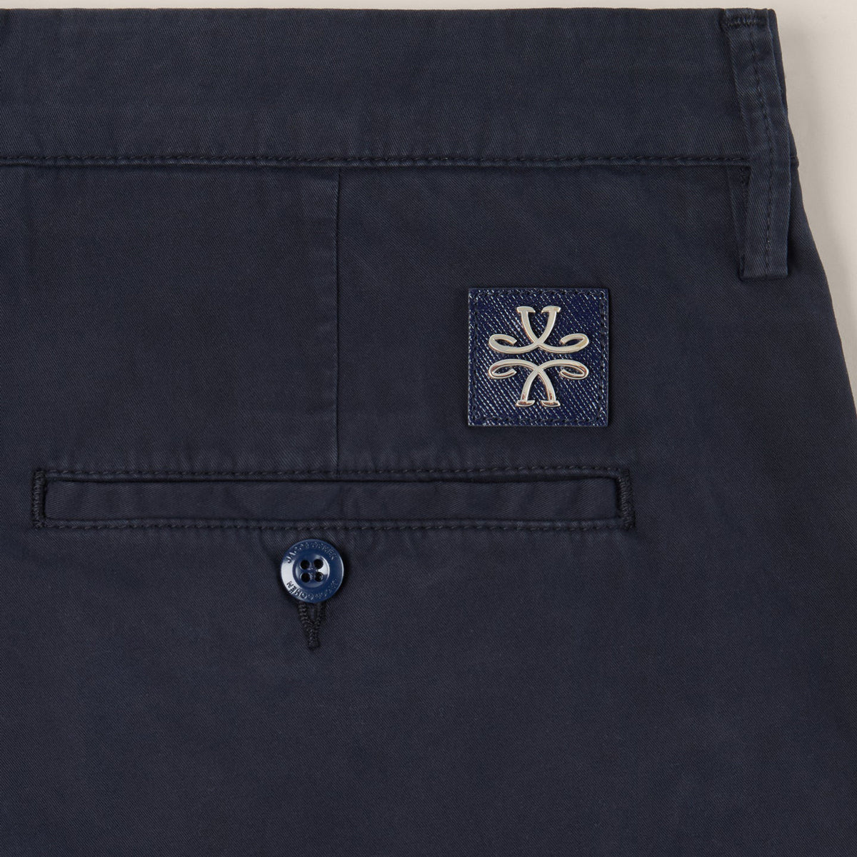Close-up of dark navy trousers showing the back waistband, a rear pocket with a button, and a square embroidered patch with a stylized logo. Jacob Cohën Chinos available at Sakks Menswear in Southampton.