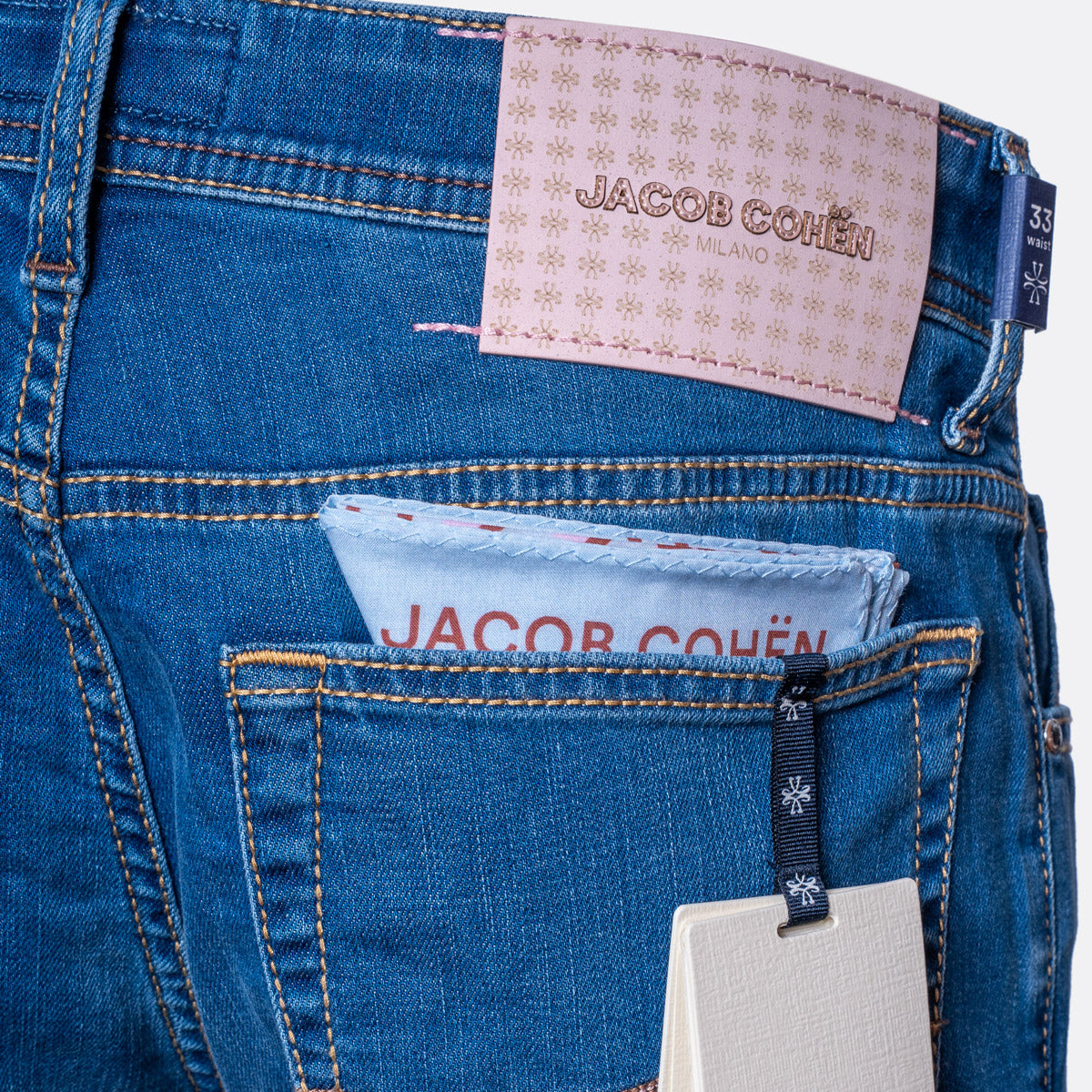 Close-up of the back waistband and pocket area of blue denim jeans with a pink Jacob Cohen label at the back waist and a light blue paper insert in the back pocket. Jacob Cohën Jeans available at Sakks Menswear in Southampton.
