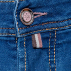 Close-up of blue denim jeans showing the button closure, contrast stitching, and a small pink, white, and gray striped fabric tab. Jacob Cohën Jeans available at Sakks Menswear in Southampton.