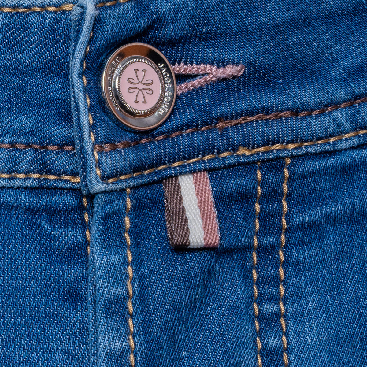 Close-up of blue denim jeans showing the button closure, contrast stitching, and a small pink, white, and gray striped fabric tab. Jacob Cohën Jeans available at Sakks Menswear in Southampton.