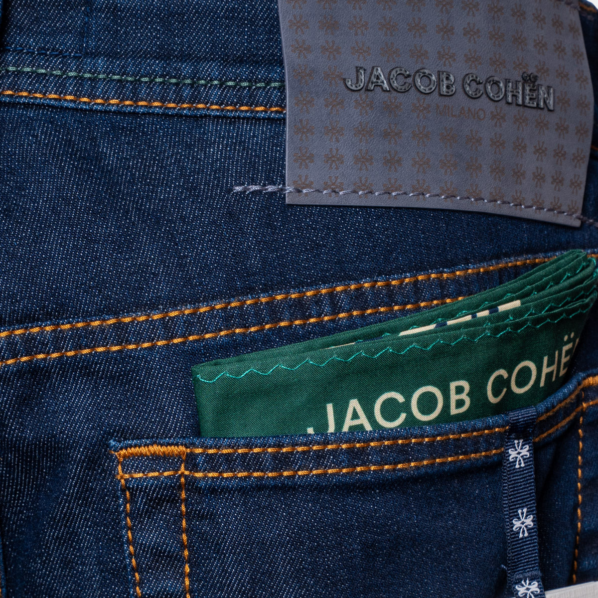 Close-up of the back pocket of dark blue denim jeans with orange contrast stitching, a green fabric pocket protector displaying 'JACOB COHEN', and a gray brand patch on the waistband. Jacob Cohën Jeans available at Sakks Menswear in Southampton.