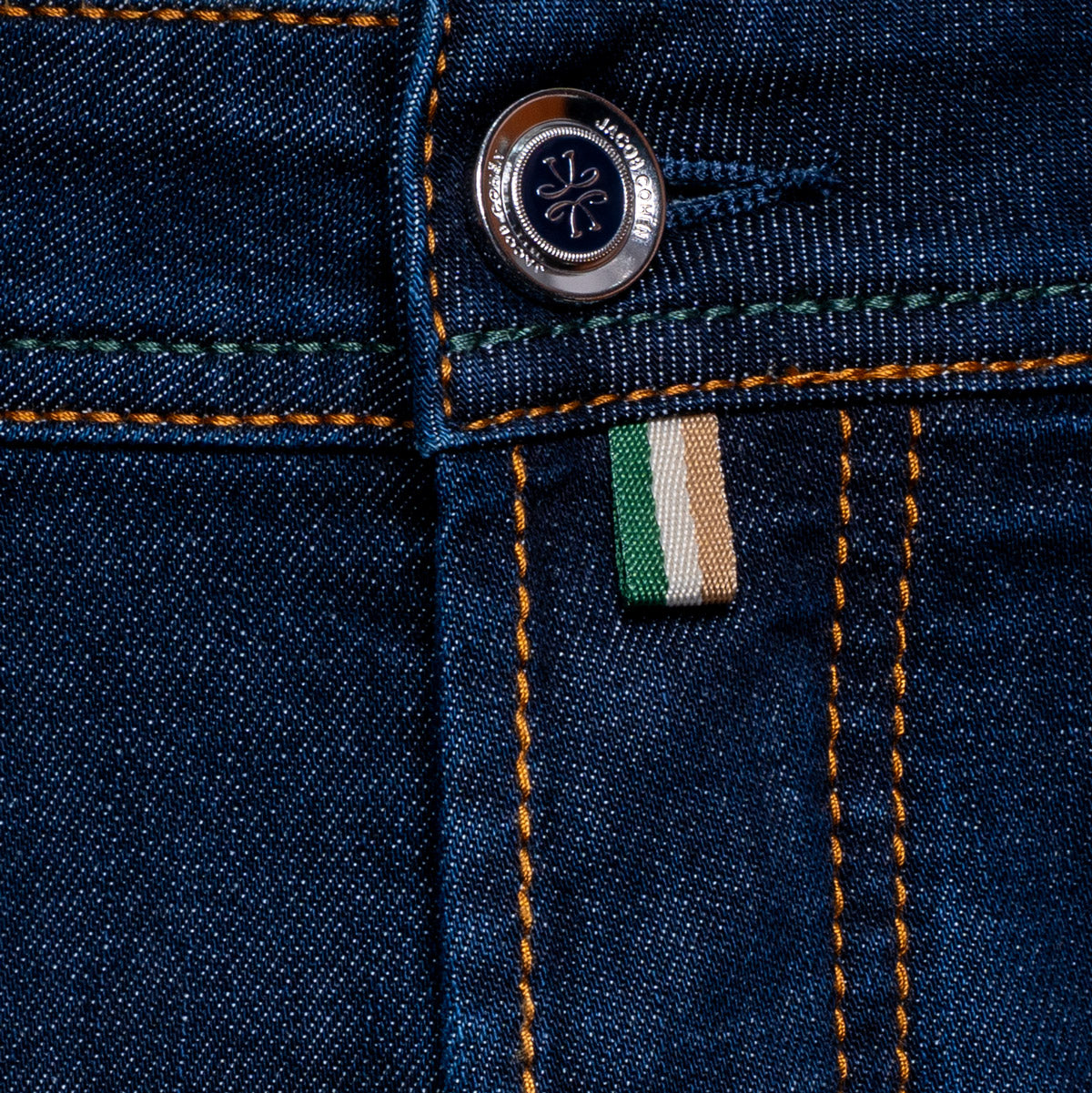 Close-up of dark blue denim jeans showing the button, orange contrast stitching, and a small green, white, and beige fabric brand tab. Jacob Cohën Jeans available at Sakks Menswear in Southampton.