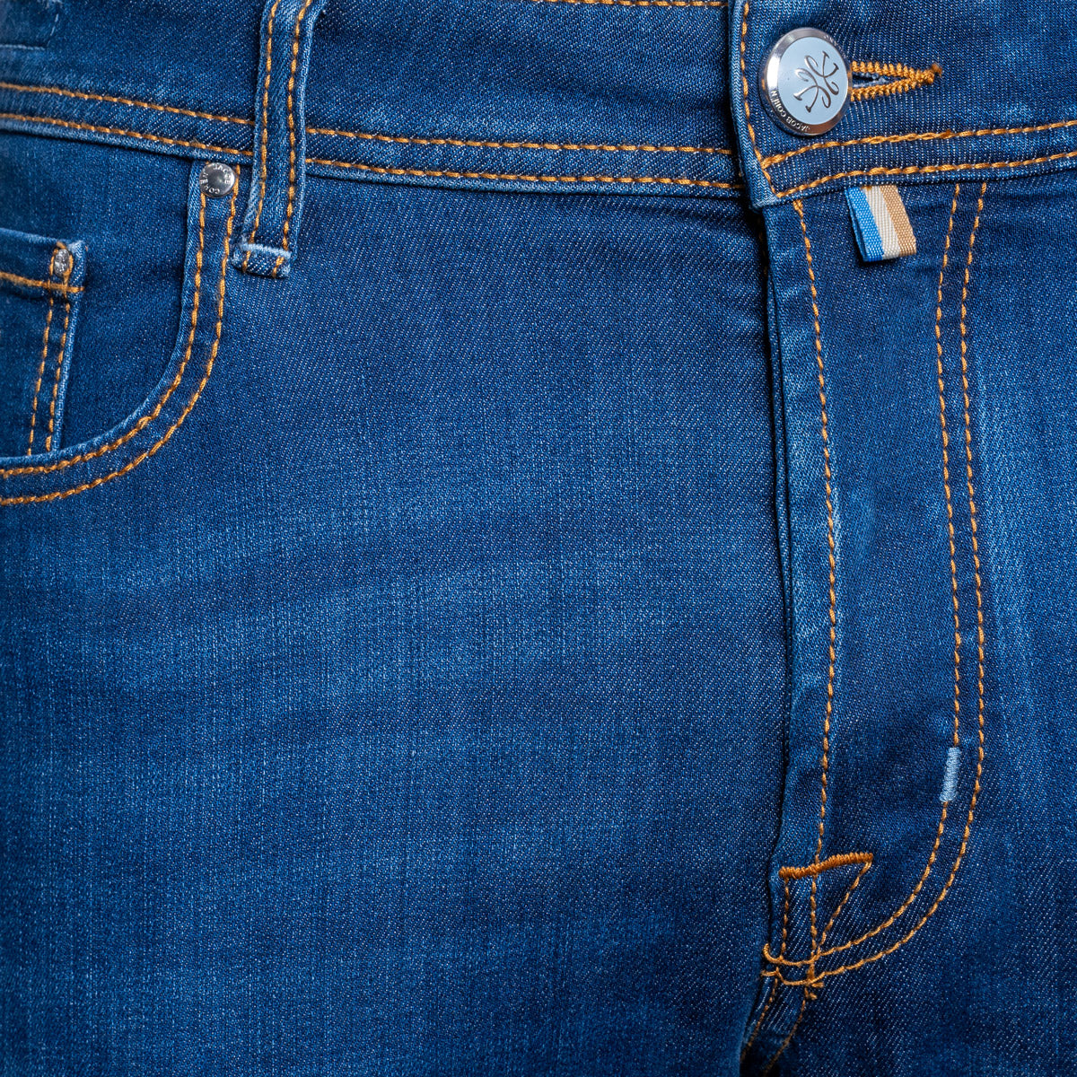 Close-up of a pair of blue denim jeans showing the waistband, button, pocket, and orange contrast stitching. Jacob Cohën Jeans available at Sakks Menswear in Southampton.
