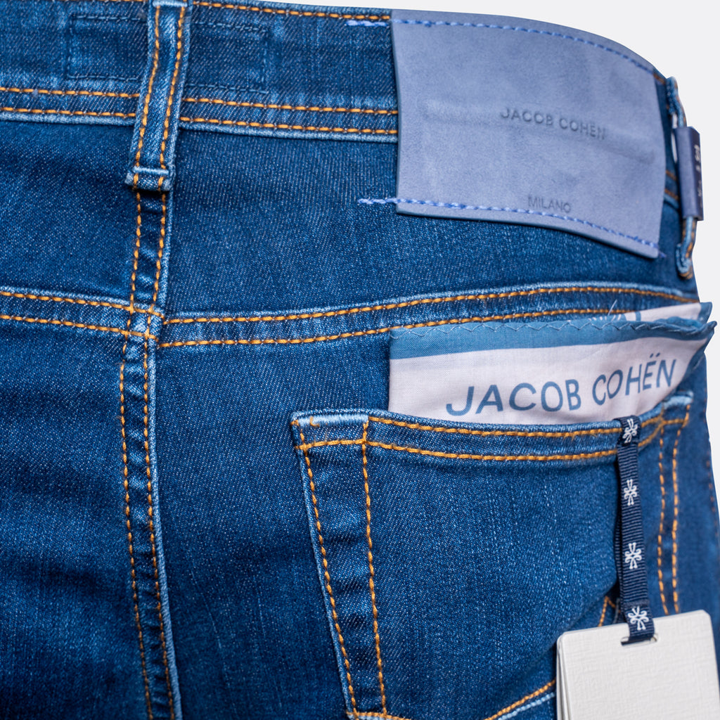 Close-up of the back waistband and pocket area of blue denim jeans by Jacob Cohen, showing orange contrast stitching, a light blue leather patch, a white brand label, and a hangtag. Jacob Cohën Jeans available at Sakks Menswear in Southampton.