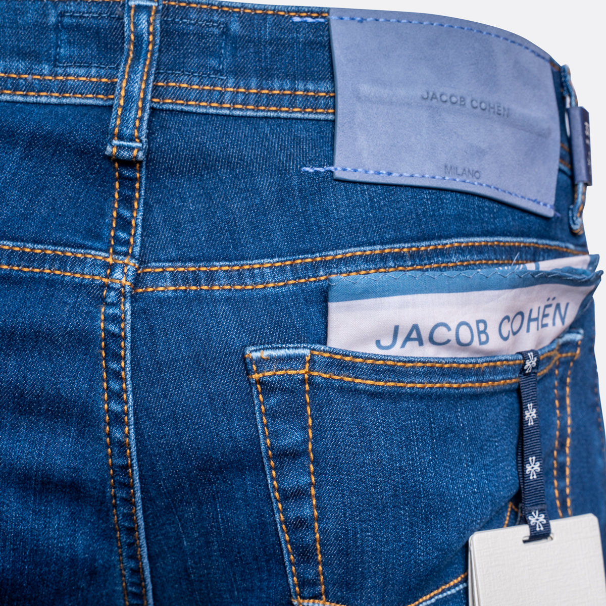Close-up of the back waistband and pocket area of blue denim jeans by Jacob Cohen, showing orange contrast stitching, a light blue leather patch, a white brand label, and a hangtag. Jacob Cohën Jeans available at Sakks Menswear in Southampton.