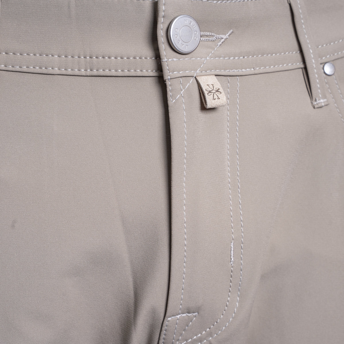 Close-up of the waistband of khaki trousers, showing a silver metal button, white contrast stitching, and a small beige fabric brand tag with a stylized logo. Jacob Cohën Chinos at Sakks Menswear in Southampton.