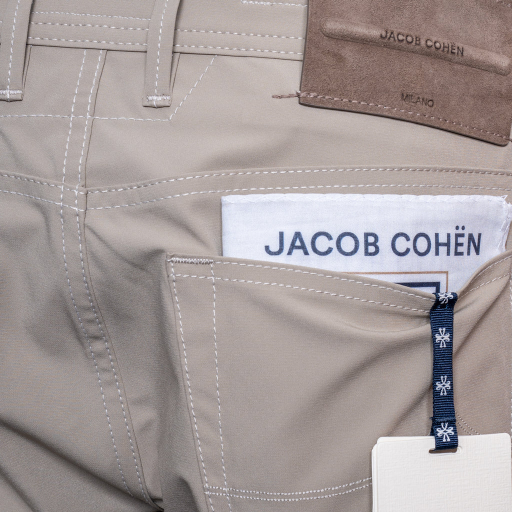 Close-up of light beige jeans showing the back pocket with a white paper tag reading JACOB COHËN, a dark blue branded zipper pull, and part of a brown leather patch on the waistband. Jacob Cohën Chinos at Sakks Menswear in Southampton.