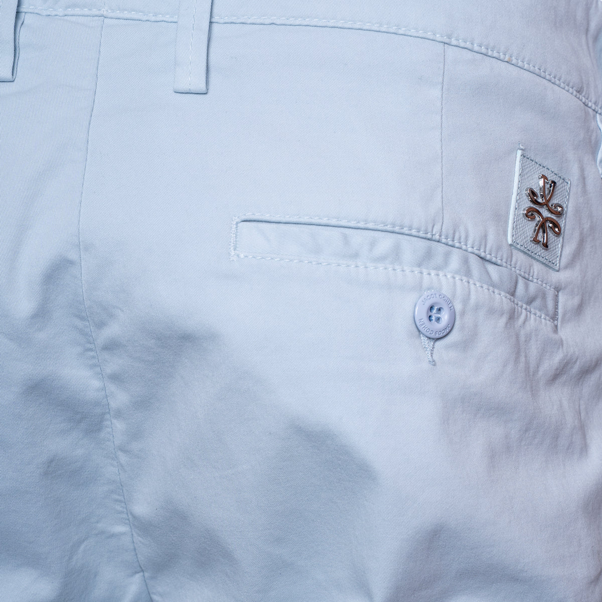 Close-up of the back waistband and pocket area of light blue shorts, showing a buttoned welt pocket, belt loops, and a small embroidered logo patch on the pocket. Jacob Cohën Shorts available at Sakks Menswear in Southampton.