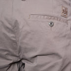 Close-up of a light taupe-gray pair of pants showing a pocket with a silver button and a small woven brand patch. Jacob Cohën Shorts available at Sakks Menswear in Southampton.