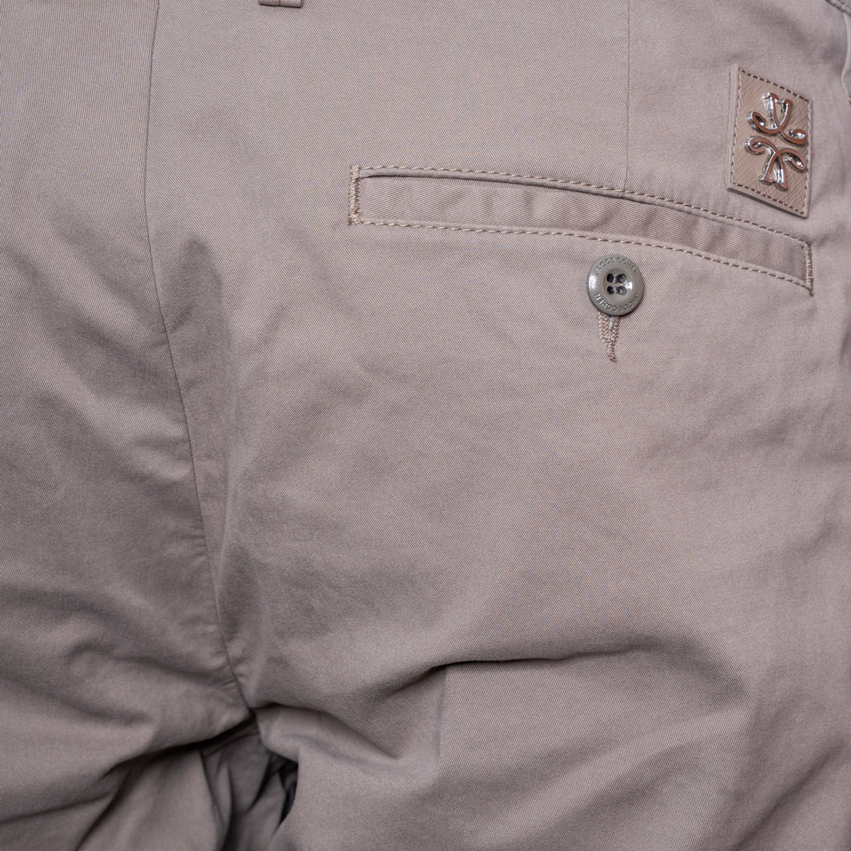 Close-up of a light taupe-gray pair of pants showing a pocket with a silver button and a small woven brand patch. Jacob Cohën Shorts available at Sakks Menswear in Southampton.
