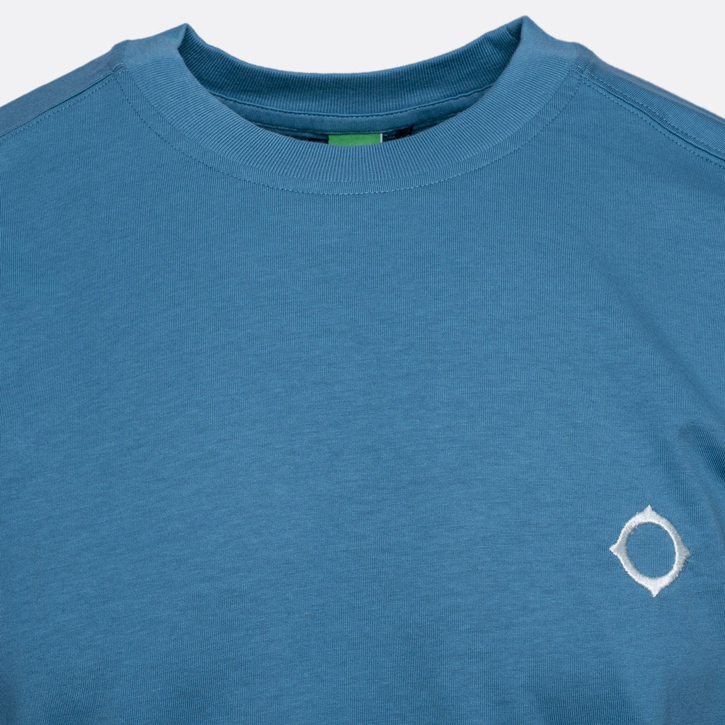 A close-up of a heathered blue crewneck t-shirt featuring a small white circular logo on the left chest, set against a light gray background. MA.Strum T-Shirts available at Sakks Menswear in Southampton.