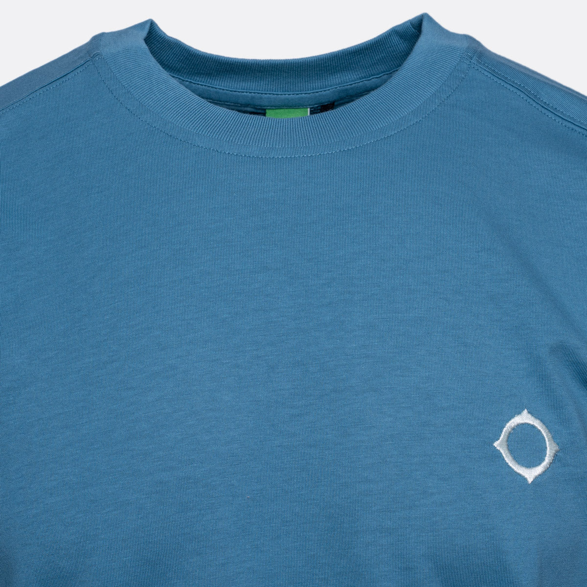 A close-up of a heathered blue crewneck t-shirt featuring a small white circular logo on the left chest, set against a light gray background. MA.Strum T-Shirts available at Sakks Menswear in Southampton.