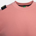 Close-up of a dusty pink crewneck sweatshirt, showing the textured fabric, a black shoulder patch with a yellow-green logo, and the inner collar. MA.Strum Sweatshirt available at Sakks Menswear in Southampton.