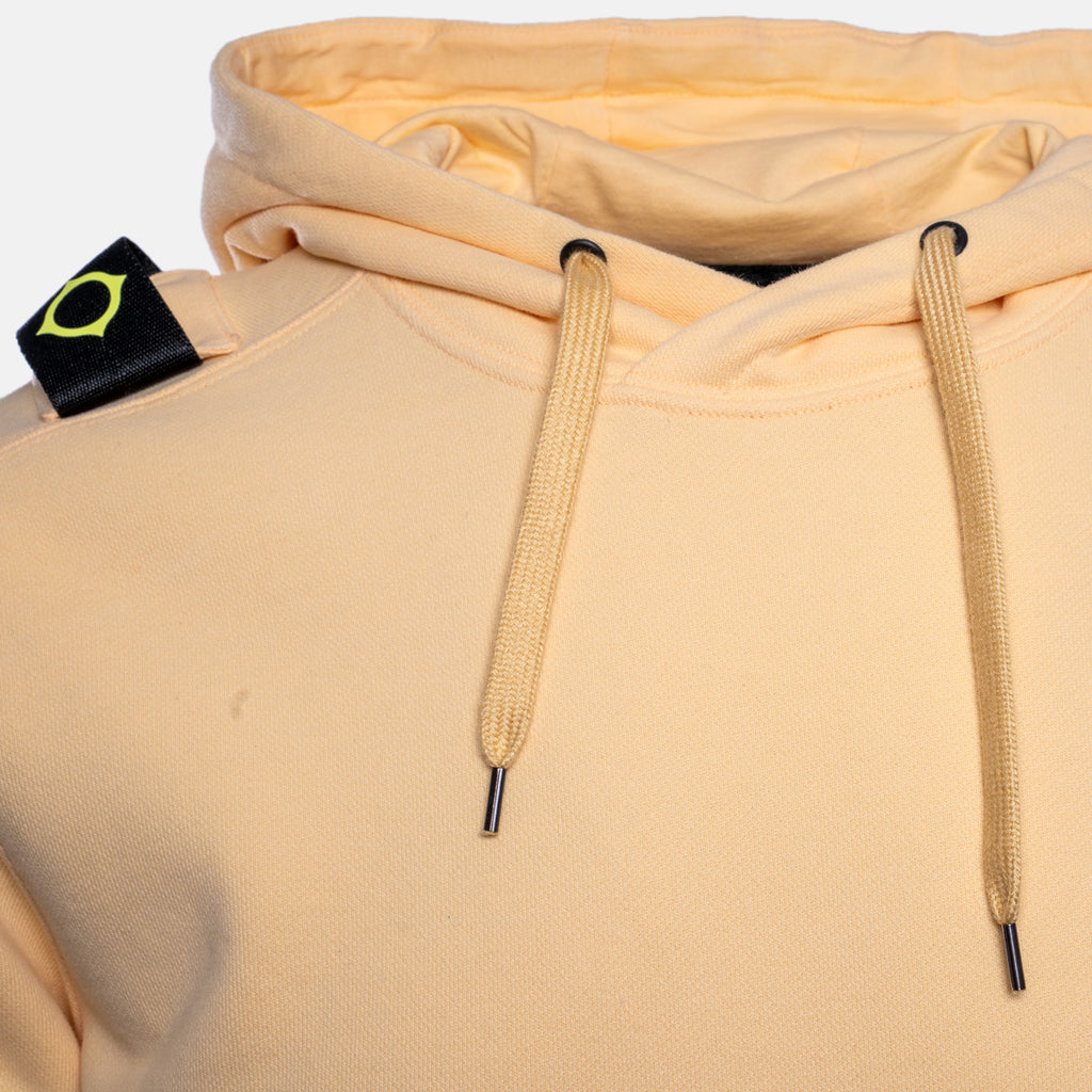 Close-up of a light peach-beige pullover hoodie with matching drawstrings and a black neck label featuring a yellow-green geometric logo. MA.Strum Hoodie available at Sakks Menswear in Southampton.