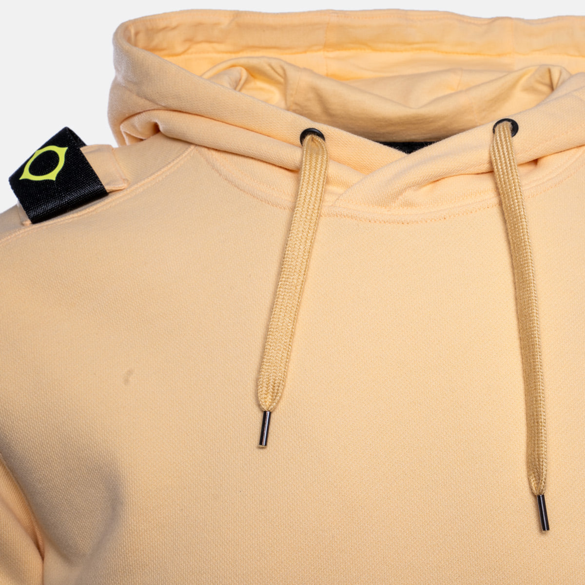 Close-up of a light peach-beige pullover hoodie with matching drawstrings and a black neck label featuring a yellow-green geometric logo. MA.Strum Hoodie available at Sakks Menswear in Southampton.